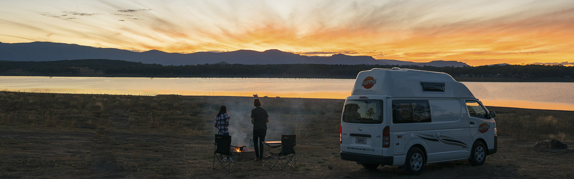 Two people watch a sunset over a lake next to their Hippie Camper